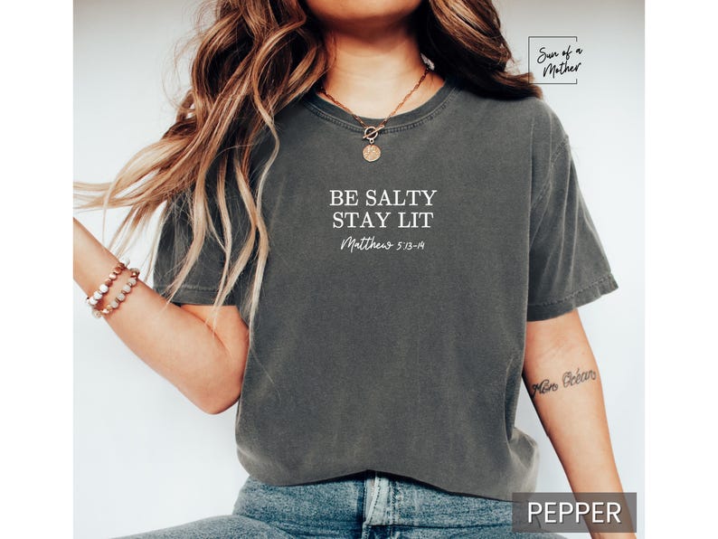 Puede incluir: Una camiseta gris oscuro con el texto "Be Salty Stay Lit" y "Matthew 5:13-14" en letras blancas.