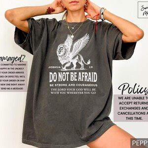 May include: Dark gray t-shirt featuring a white illustration of a lion with wings, accompanied by the text "DO NOT BE AFRAID" and a Bible verse. The shirt has a relaxed fit and is made of a soft material.