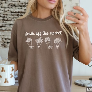 May include: Espresso-colored t-shirt with the text "fresh off the market" above four bouquets of flowers. A white mug with teddy bear designs is held in the left hand. The shirt is a neutral color.