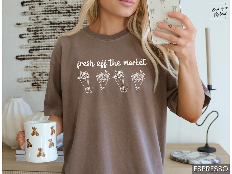 May include: Espresso-colored t-shirt with the text "fresh off the market" above four bouquets of flowers. A white mug with teddy bear designs is held in the left hand. The shirt is a neutral color.