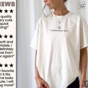 May include: A white t-shirt with a graphic of a potted plant and the text "I'd Propagate That". The shirt is shown on a model and the color is ivory.