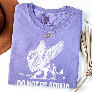 May include: A lilac-colored t-shirt with a white graphic of a winged lion and the text "Joshua 1:9 DO NOT BE AFRAID." A gold necklace with a circular pendant rests on the shirt. A brown hat and dried palm leaf are also in the image.