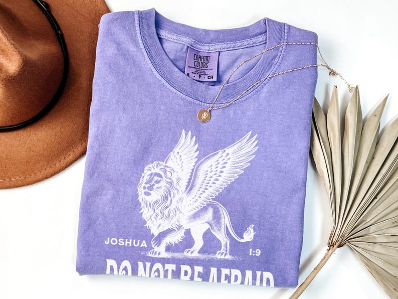 May include: A lilac-colored t-shirt with a white graphic of a winged lion and the text "Joshua 1:9 DO NOT BE AFRAID." A gold necklace with a circular pendant rests on the shirt. A brown hat and dried palm leaf are also in the image.