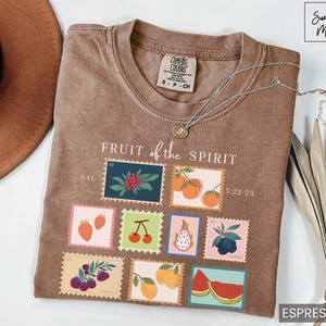 May include: A brown t-shirt with the text "FRUIT of the SPIRIT" and a graphic of nine postage stamps featuring different fruits. The stamps are labeled with the text "GAL" and "5:22-23".