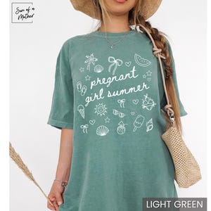May include: Light green t-shirt with the text "pregnant girl summer" and summer-themed graphics in white. The shirt has a relaxed fit and short sleeves. The image also shows a straw hat and a woven bag.