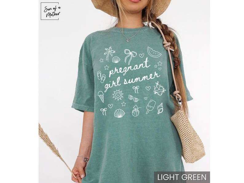 May include: Light green t-shirt with the text "pregnant girl summer" and summer-themed graphics in white. The shirt has a relaxed fit and short sleeves. The image also shows a straw hat and a woven bag.