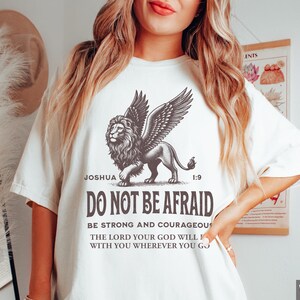 May include: White t-shirt featuring a detailed illustration of a winged lion with the text "DO NOT BE AFRAID" and a Bible verse. The shirt's design includes the word "Joshua" and the number "1:9".