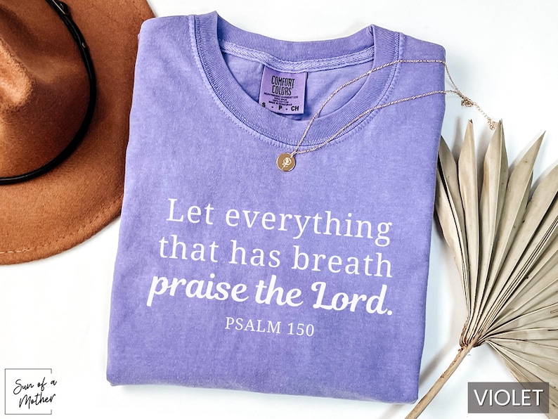 May include: A violet-colored t-shirt with the text "Let everything that has breath praise the Lord. Psalm 150" in white. A gold necklace and a brown hat are also visible. The shirt is part of a clothing collection.