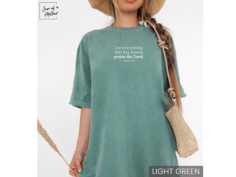 May include: Light green t-shirt with the text "Let everything that has breath praise the Lord. Psalm 136" in white. The shirt is a solid color and has short sleeves. The image also shows a straw hat and a woven bag.