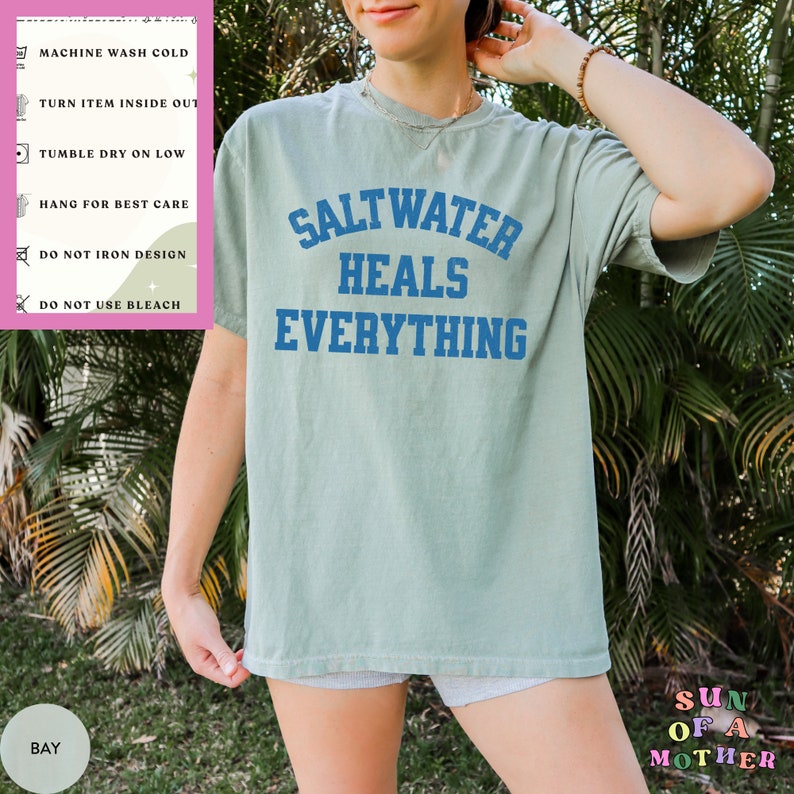 May include: A light blue t-shirt with the text "Saltwater Heals Everything" in blue lettering. The shirt is worn by a person standing in front of green foliage. The care instructions are visible on a pink label.
