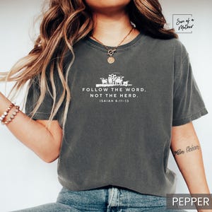Peut inclure: Un t-shirt gris fonc&eacute; avec un motif blanc repr&eacute;sentant un groupe de moutons et le texte "FOLLOW THE WORD, NOT THE HERD. ISAIAH 8:11-13".