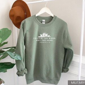 May include: Military green sweatshirt with the text "FOLLOW THE WORD, NOT THE HERD." and Isaiah 8:11-13 printed in white. A brown hat hangs on a white rack behind the sweatshirt.