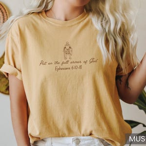 Puede incluir: Camiseta amarilla mostaza con un dibujo lineal de una persona con armadura y el texto "Ponte la armadura completa de Dios Efesios 6:10-18".