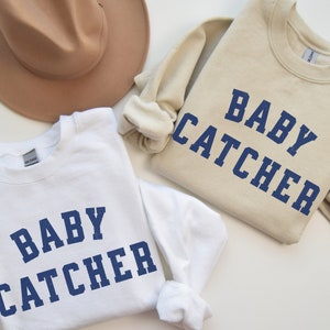 May include: Two white sweatshirts with the text "Baby Catcher" in blue lettering. One sweatshirt is beige and the other is white.