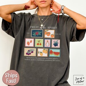May include: A dark gray t-shirt with a graphic print of fruit and the text "FRUIT of the SPIRIT GAL 5:22-23" and "But the fruit of the spirit is love, joy, peace, patience, kindness, goodness, faithfulness, gentleness, self control".