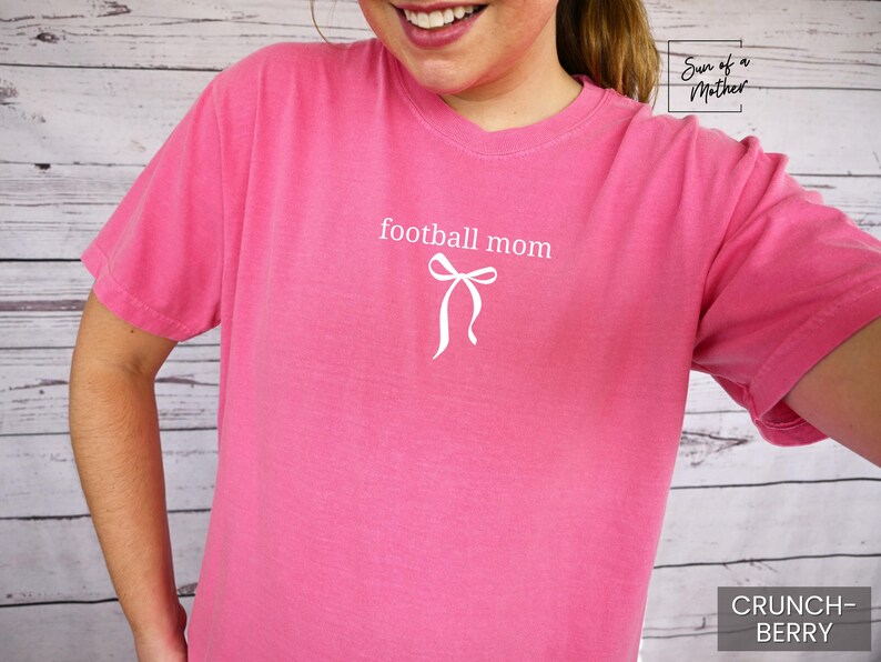 May include: A pink t-shirt with the words "football mom" and a white bow graphic. The shirt is a solid color and has a crew neck. The text "Crunch Berry" is in the bottom right corner.