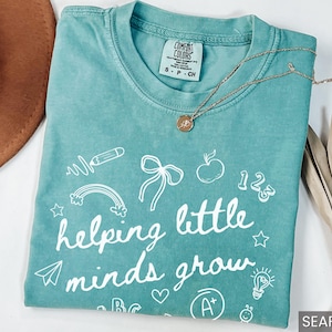 May include: Seafoam green t-shirt with white text that reads "helping little minds grow". The shirt is decorated with various school-themed illustrations, including a pencil, rainbow, apple, and a lightbulb.