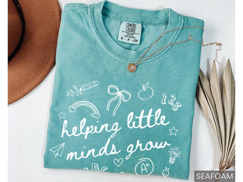 May include: Seafoam green t-shirt with white text that reads "helping little minds grow". The shirt is decorated with various school-themed illustrations, including a pencil, rainbow, apple, and a lightbulb.