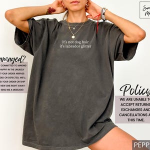 May include: Dark gray t-shirt with the text "it's not dog hair it's labrador glitter" in white. The shirt has a relaxed fit. Additional text on the image includes "Damaged?" and "Policy".