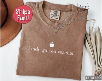 以下が含まれることがあります： 白いリンゴのグラフィックと「kindergarten teacher」の文字が書かれた茶色のTシャツ。このTシャツは柔らかく快適な素材で作られています。