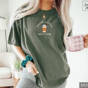 May include: Moss green t-shirt with a graphic of a pumpkin spice latte and the text "feeling a little spicy today." A pink mug with "Cool Mom Club" is held in the hand. The shirt is a relaxed fit.