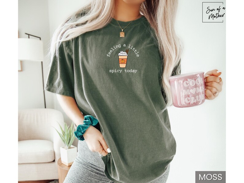May include: Moss green t-shirt with a graphic of a pumpkin spice latte and the text "feeling a little spicy today." A pink mug with "Cool Mom Club" is held in the hand. The shirt is a relaxed fit.