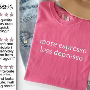 Op de afbeelding: Een Crunch Berry gekleurd t-shirt met de witte tekst "more espresso less depresso". Het shirt is opgevouwen en op een spijkerbroek gelegd. Een beige hoed is zichtbaar op de achtergrond.