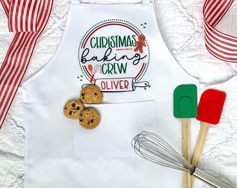 May include: White apron with the text "Christmas Baking Crew" and the name "Oliver" in a circle design. The apron has a pocket with chocolate chip cookies. A green and red spatula, a whisk, and a red and white striped ribbon are also in the image.