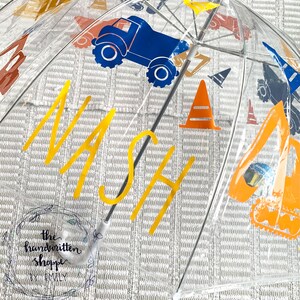 May include: Clear umbrella with construction vehicle graphics in blue, orange, and brown. The name "NASH" is printed in yellow. The umbrella also features an orange traffic cone design. The handwritten shoppe by Emily logo is visible.