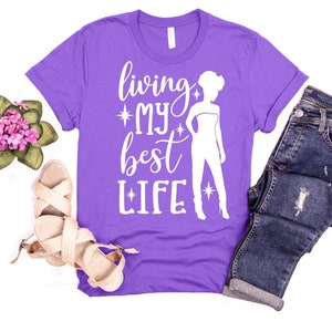 May include: Purple t-shirt with a white silhouette of a woman and the text "living my best life" with stars on either side of the words "my" and "life".