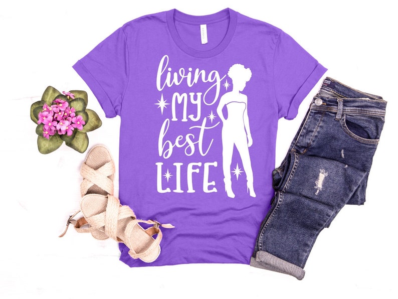 May include: Purple t-shirt with a white silhouette of a woman and the text "living my best life" with stars on either side of the words "my" and "life".