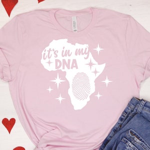 May include: Pink t-shirt with a white graphic of the continent of Africa with a fingerprint inside and the text "it's in my DNA" above it. There are white stars around the graphic.