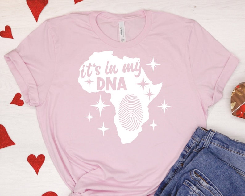 May include: Pink t-shirt with a white graphic of the continent of Africa with a fingerprint inside and the text "it's in my DNA" above it. There are white stars around the graphic.