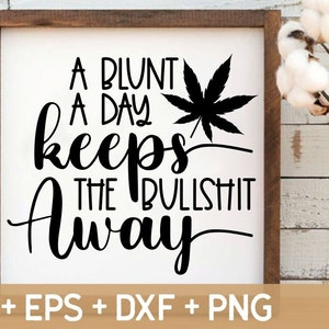May include: A framed sign with the text "A BLUNT A DAY keeps THE BULLSHIT Away" in black script. A black cannabis leaf is next to the text. The sign is on a white background with a wooden frame.