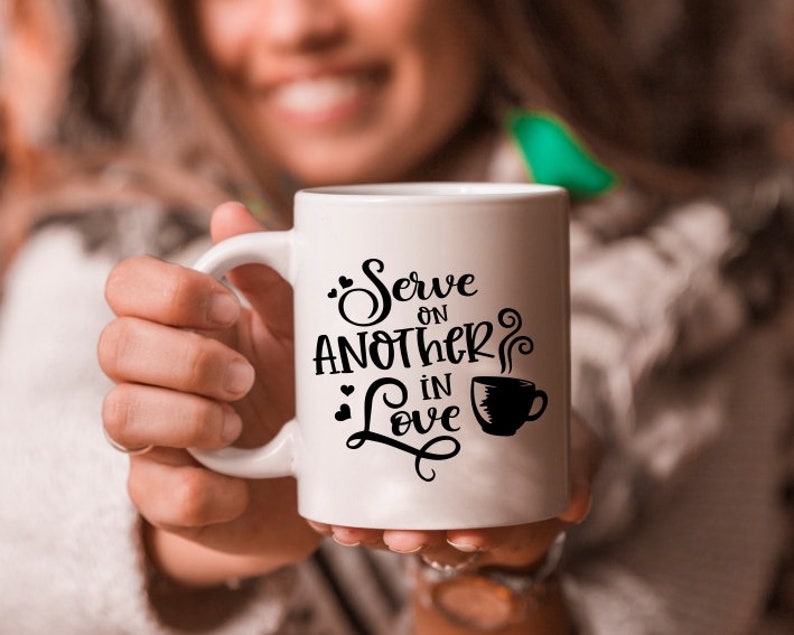 Puede incluir: Taza de cer&aacute;mica blanca con texto negro que dice "Serve on another in love" con un peque&ntilde;o gr&aacute;fico de taza de caf&eacute; negro.