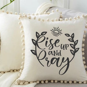 Puede incluir: Almohada blanca con un dise&ntilde;o negro que dice "Rise up and pray" con un sol y hojas alrededor del texto. La almohada tiene un ribete de pompones blancos.