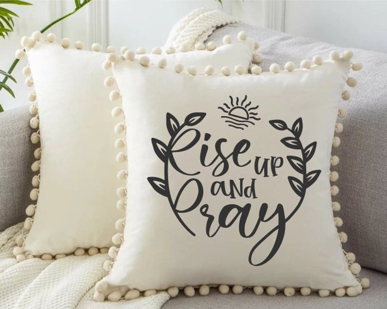 Puede incluir: Almohada blanca con un dise&ntilde;o negro que dice "Rise up and pray" con un sol y hojas alrededor del texto. La almohada tiene un ribete de pompones blancos.