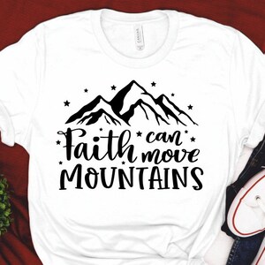 Puede incluir: Camiseta blanca con un gr&aacute;fico de una cadena monta&ntilde;osa negra y el texto "Faith can move mountains".