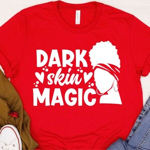 May include: Red t-shirt with a white graphic design featuring a woman's silhouette with a headscarf and the text "DARK skin MAGIC" with hearts.