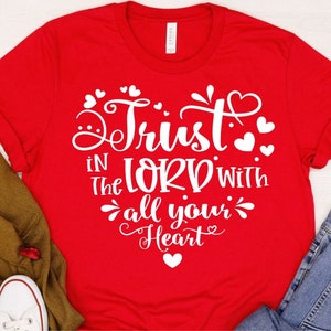 Puede incluir: Camiseta roja con texto blanco que dice "Trust in the Lord with all your heart" rodeado de peque&ntilde;os corazones.