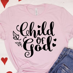 Puede incluir: Una camiseta rosa con el texto "Child of God" en letras cursivas negras. El texto est&aacute; rodeado de peque&ntilde;os corazones y hojas negras.