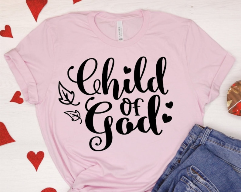 Puede incluir: Una camiseta rosa con el texto "Child of God" en letras cursivas negras. El texto est&aacute; rodeado de peque&ntilde;os corazones y hojas negras.
