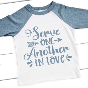 Puede incluir: Una camiseta blanca de manga larga con mangas ragl&aacute;n azules. La camiseta tiene un gr&aacute;fico azul que dice "Servir al pr&oacute;jimo con amor" con una flecha apuntando hacia la derecha.
