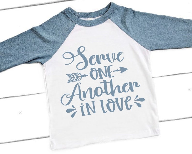 Puede incluir: Una camiseta blanca de manga larga con mangas ragl&aacute;n azules. La camiseta tiene un gr&aacute;fico azul que dice "Servir al pr&oacute;jimo con amor" con una flecha apuntando hacia la derecha.