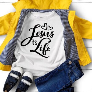 Puede incluir: Camiseta blanca de manga larga con texto negro que dice "Jesus is Life" y dos peque&ntilde;os corazones sobre el texto. La camiseta est&aacute; colocada sobre una superficie blanca con una chaqueta amarilla, unos vaqueros azules y unas zapatillas negras.