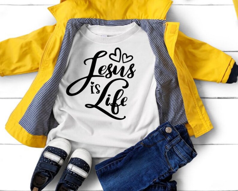 Puede incluir: Camiseta blanca de manga larga con texto negro que dice "Jesus is Life" y dos peque&ntilde;os corazones sobre el texto. La camiseta est&aacute; colocada sobre una superficie blanca con una chaqueta amarilla, unos vaqueros azules y unas zapatillas negras.