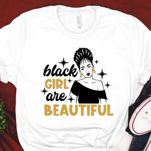 May include: White t-shirt with a black silhouette of a woman with a gold crown and the text "black girl are beautiful" in gold letters.