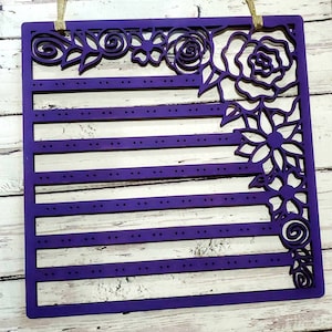 May include: Purple wooden earring holder with a floral design. The holder has six rows of holes for earrings and a floral design on the right side.