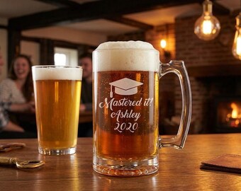 May include: A clear glass beer mug with a handle, filled with amber liquid and white foam. The front is etched with a graduation cap icon and the text "Mastered it! Ashley 2020". A second glass of beer sits to the left on a wooden surface.
