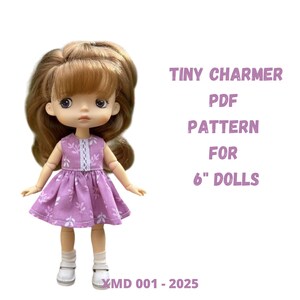 Puede incluir: Una muñeca con cabello castaño claro que lleva un vestido lavanda con detalles florales blancos y zapatos blancos. El texto a la derecha dice "TINY CHARMER PDF PATTERN FOR 6" DOLLS". La muñeca está sobre un fondo blanco.